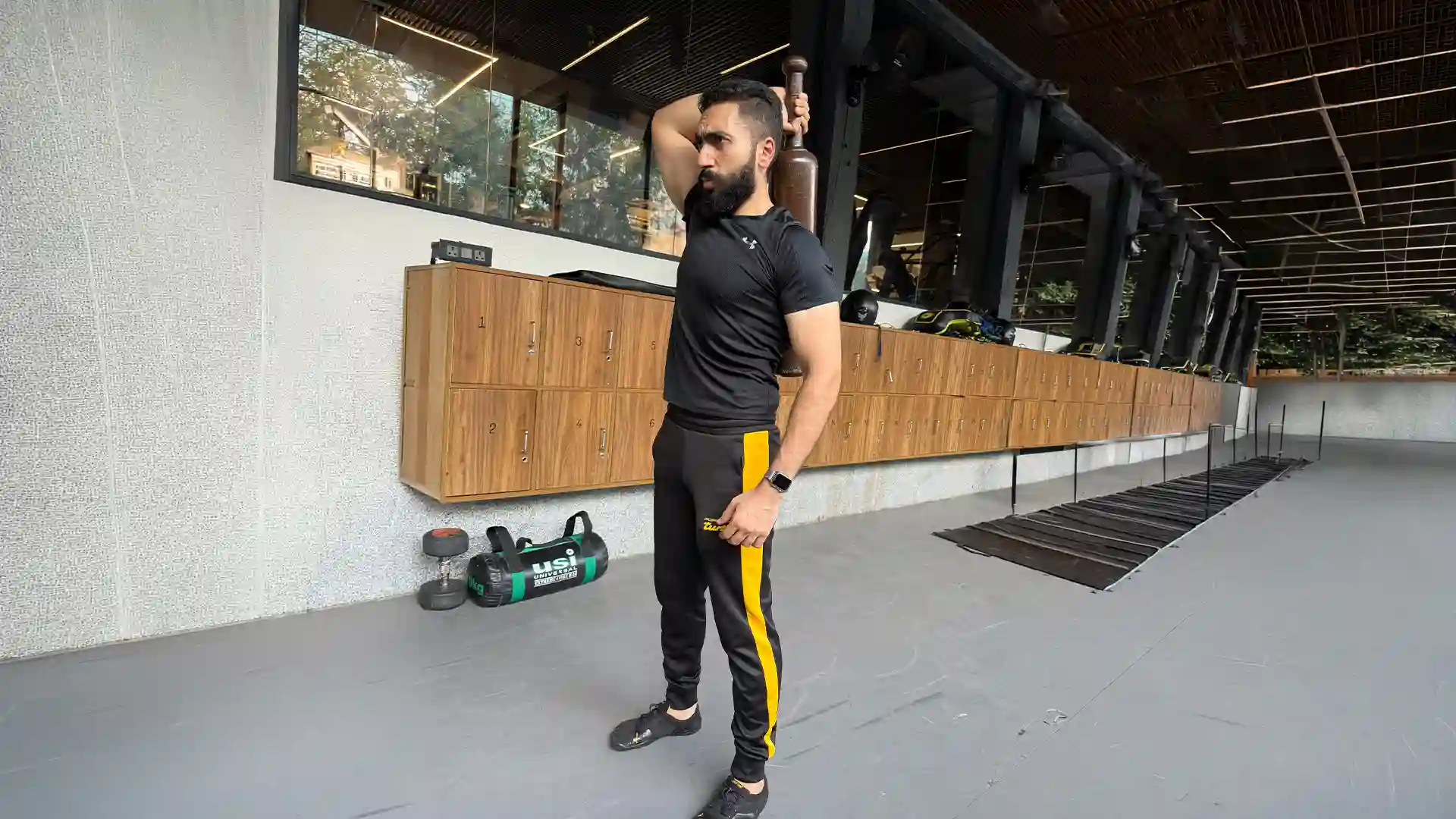A fit man with a beard, wearing a black athletic t-shirt and black track pants with a yellow stripe, performs a strength exercise using a traditional wooden Indian club (Mudgar) in a modern gym. This form of resistance exercise highlights how bodybuilding for diabetes helps build muscle mass, improve insulin sensitivity, and support metabolic health.